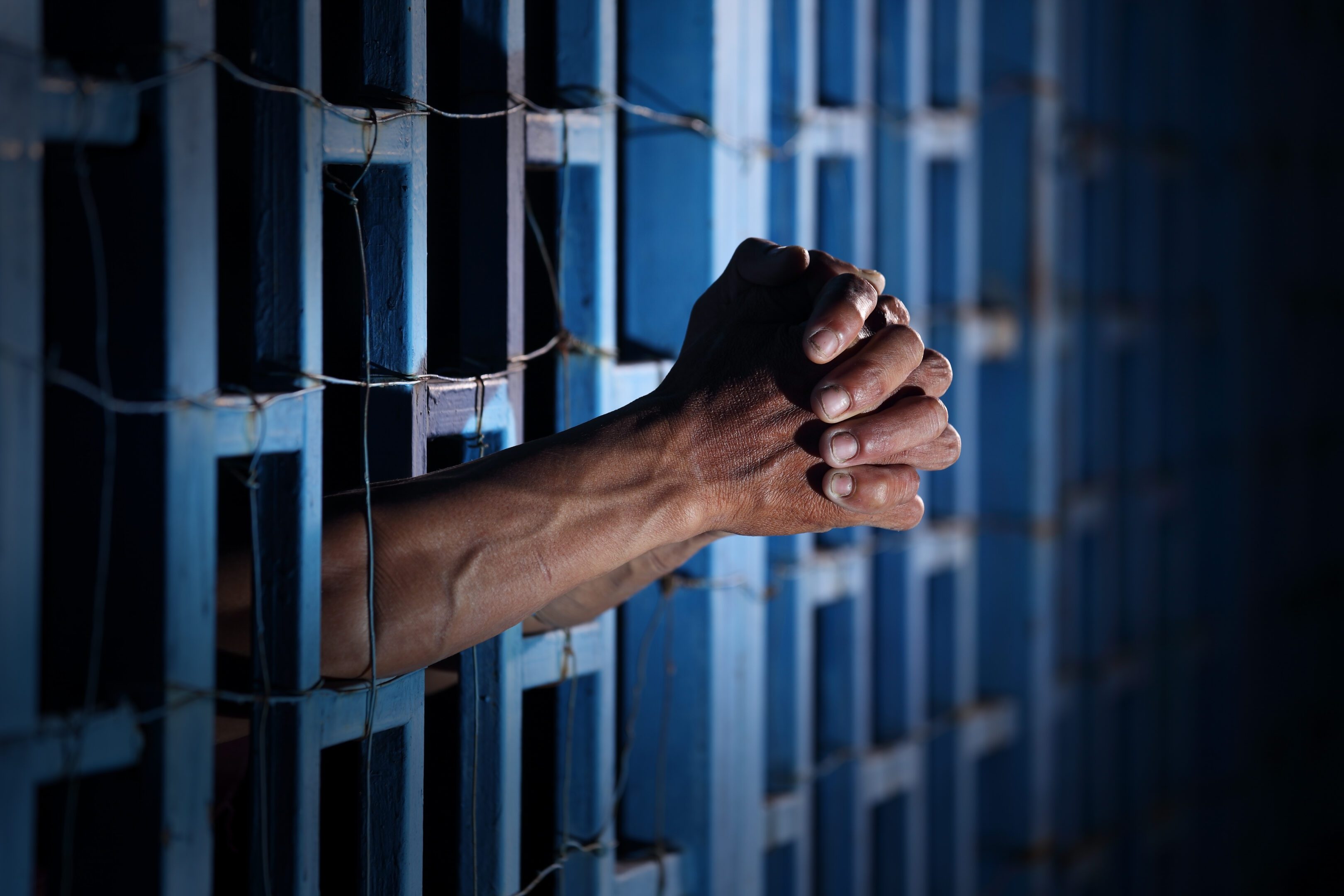 A powerful image of hands clasped together through blue prison bars, illuminated by dim light. This evokes emotions of confinement, struggle, and hope.