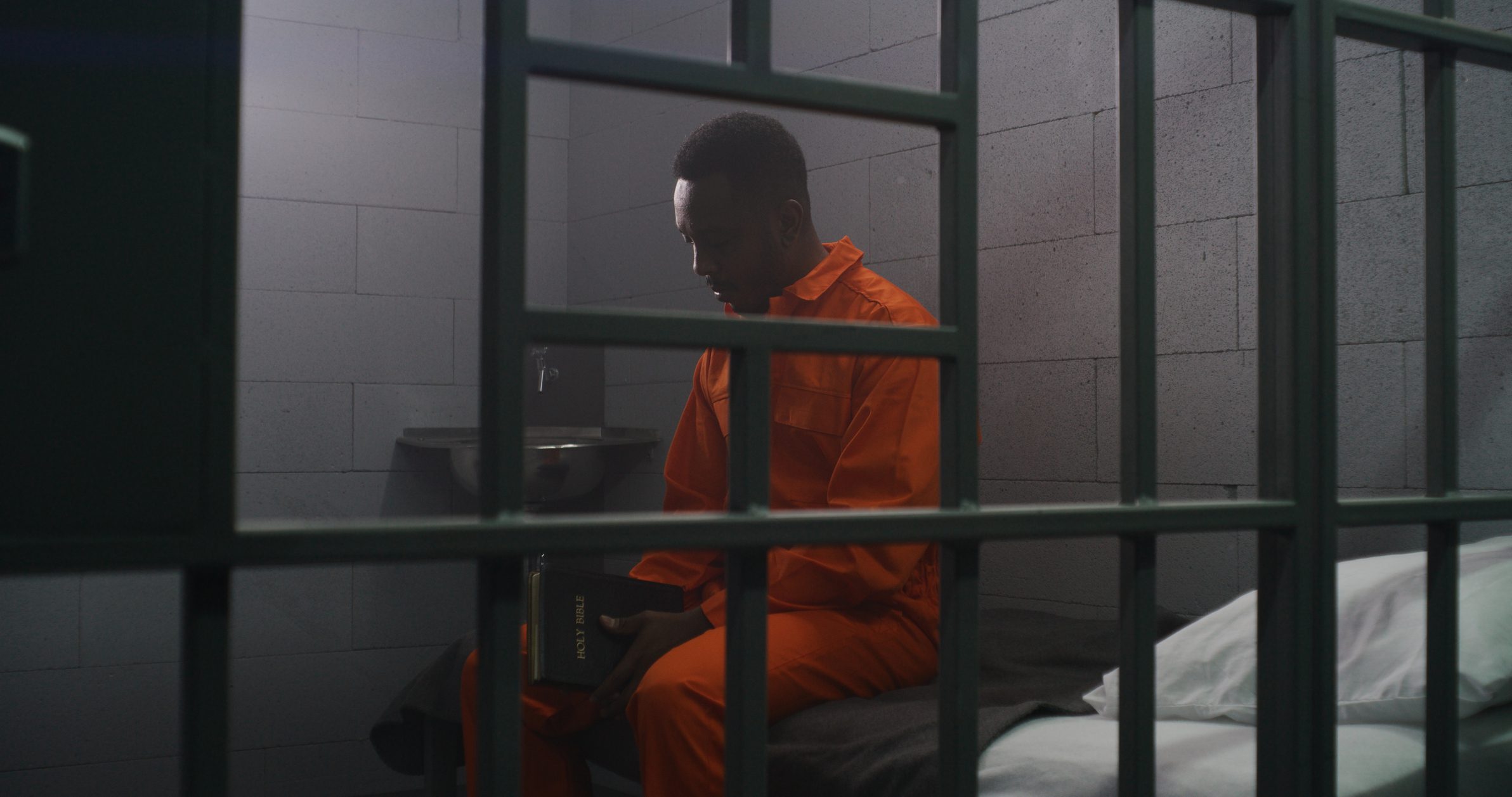 African American prisoner in orange uniform sits on the bed behind bars, reads Bible in prison cell. Criminal serves imprisonment term for crime in jail. Detention center or correctional facility.