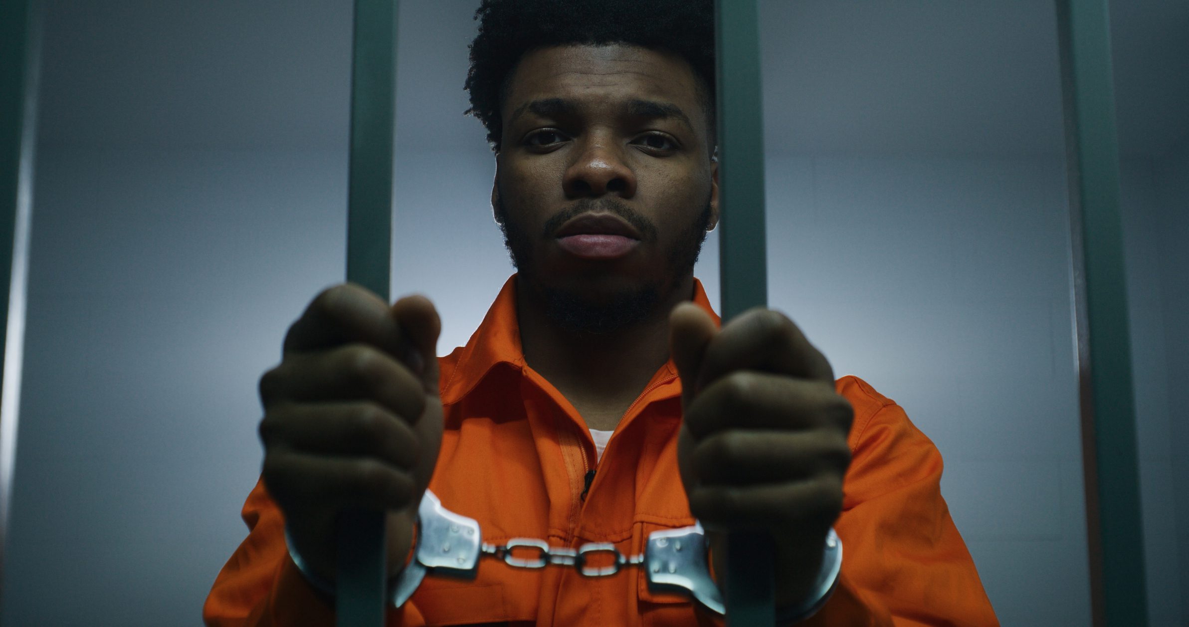 African American man in orange uniform keeps hands in handcuffs on jail cell bars. Depressed murderer serves imprisonment term in prison. Guilty prisoner in correctional facility or detention center.