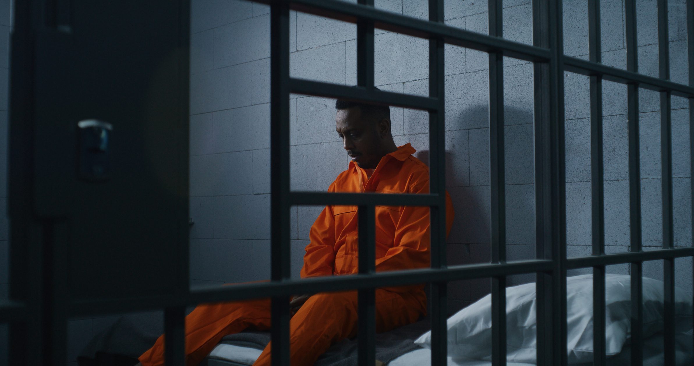 Guilty African American man in orange uniform sits on prison bed and thinks about freedom. Gloomy criminal in correctional facility or detention center. Prisoner serves imprisonment term in jail cell.