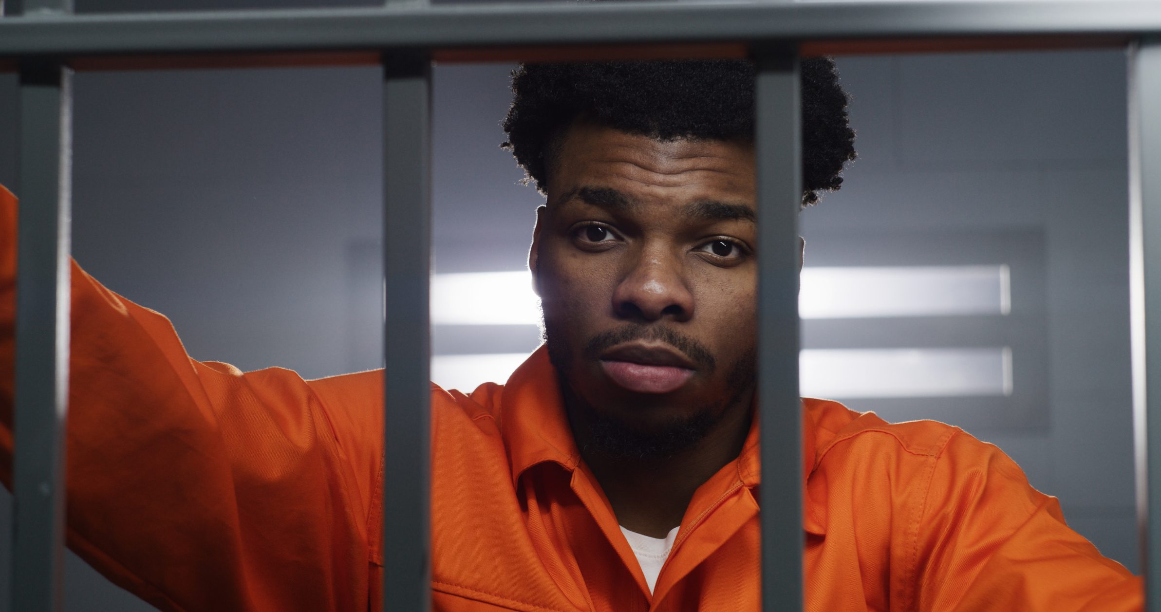 African American man in orange uniform looks at camera and kneads neck in prison cell. Prisoner serves imprisonment term in jail. Criminal in correctional facility or detention center. Portrait view.