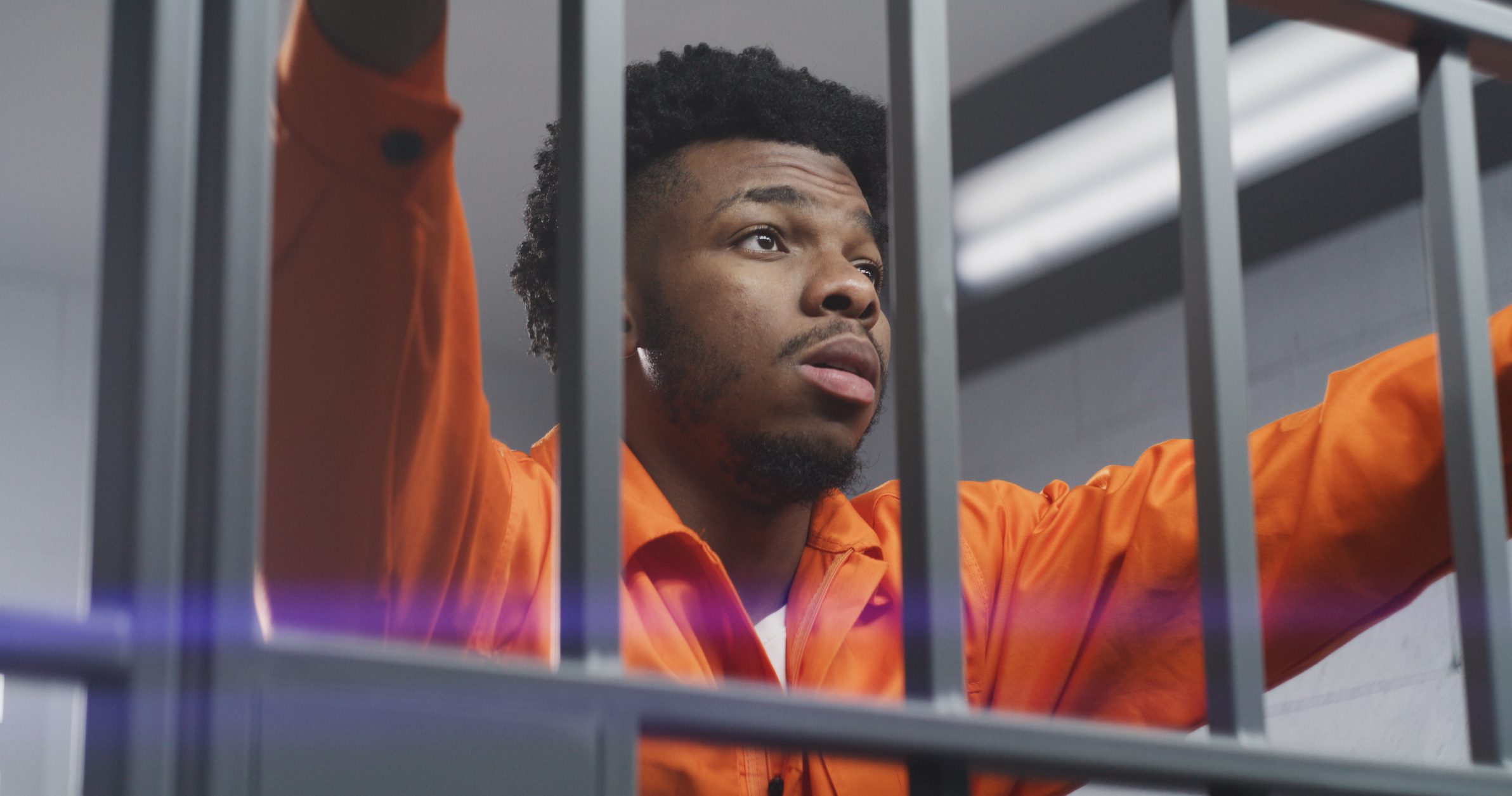 African American man in orange uniform leans hands on prison cell bars and kneads neck. Depressed prisoner serves imprisonment term in jail. Murderer in correctional facility or detention center.