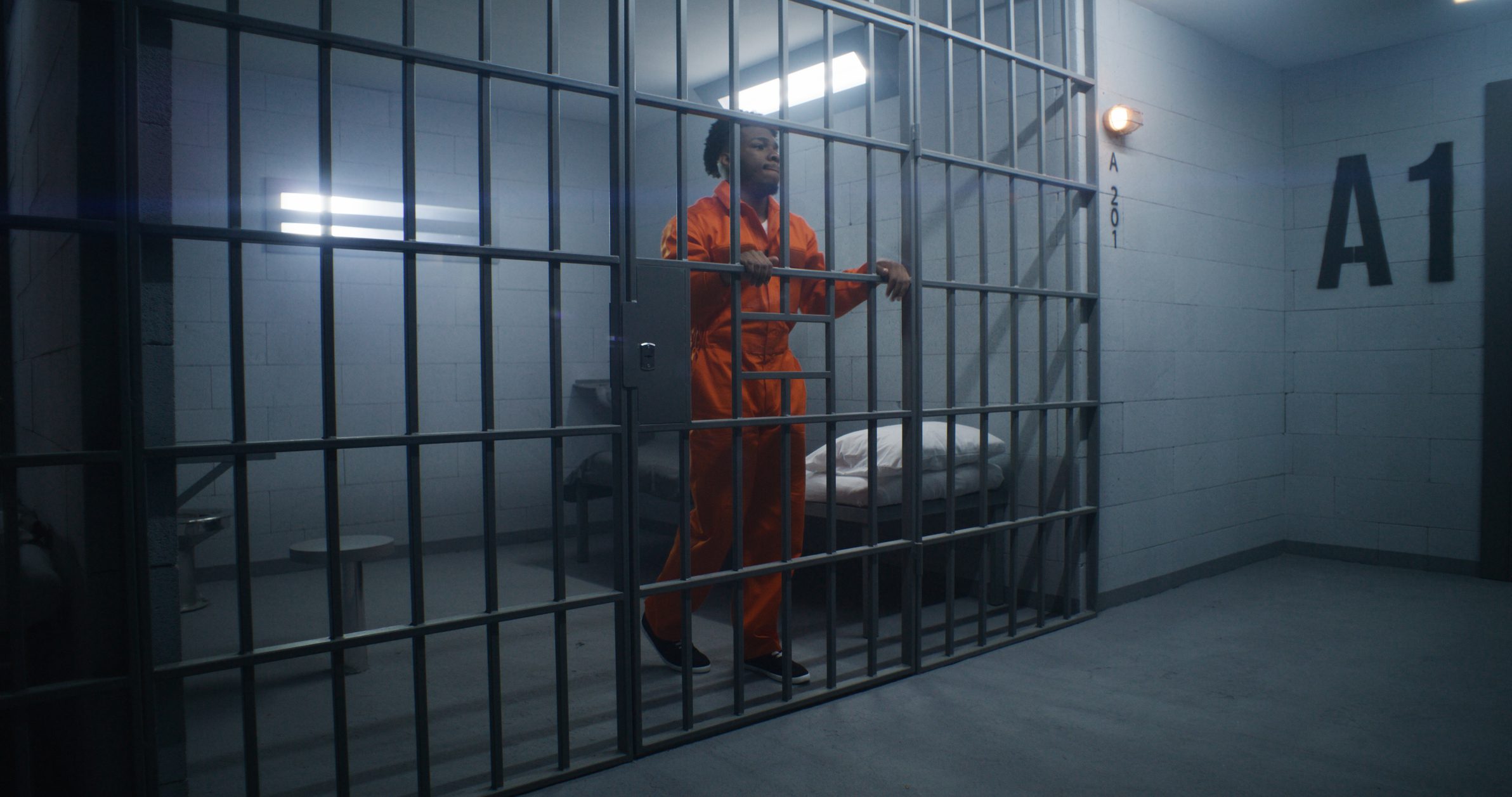 African American prisoner in orange uniform leans on jail cell bars and looks around. Guilty gangster serves imprisonment term in correctional facility. Gloomy inmate in detention center.