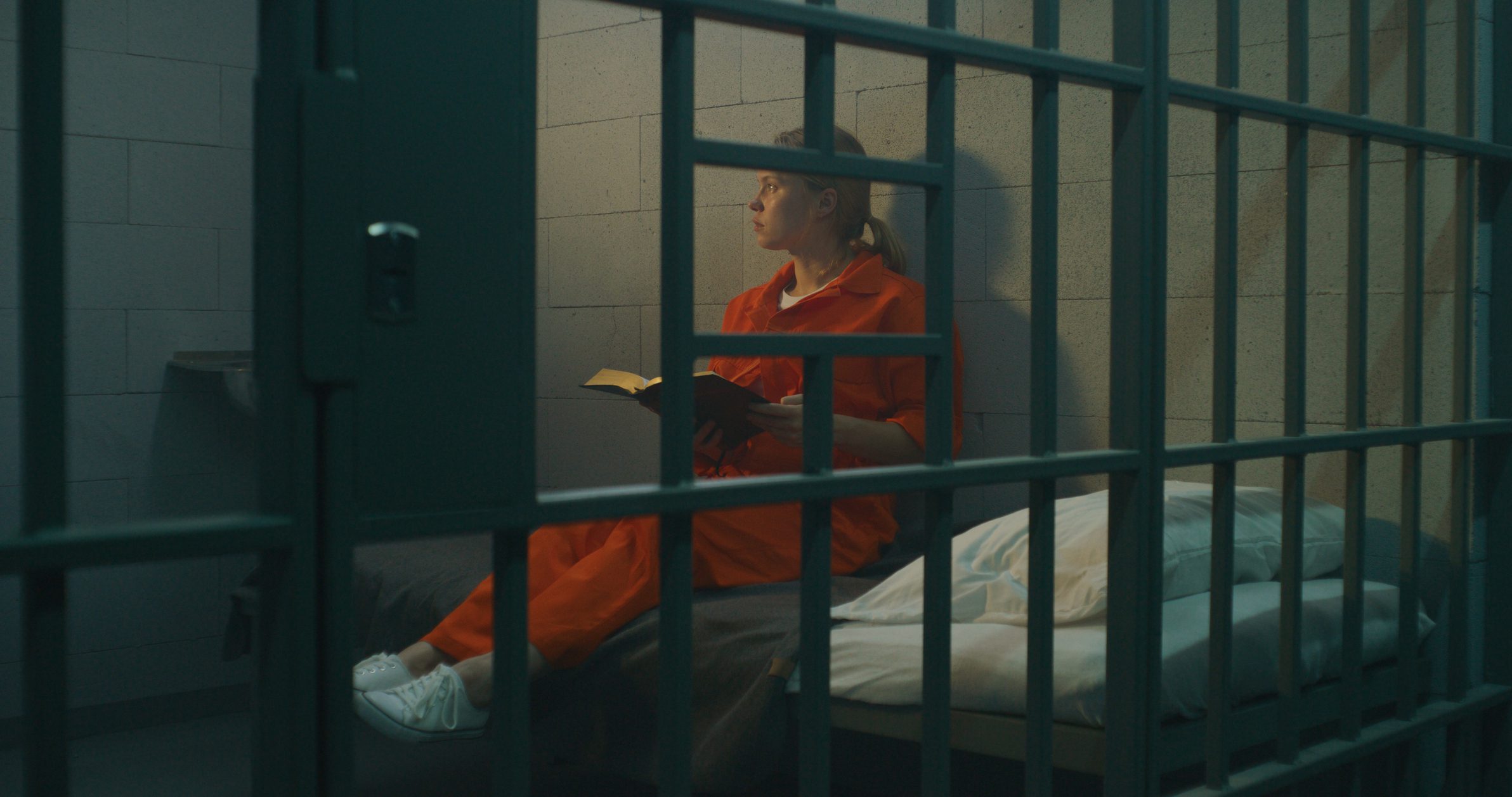 Female prisoner in orange uniform sits on bed behind metal bars, reads Bible in prison cell, looks at barred window. Woman criminal serves imprisonment term for crime in jail or correctional facility.