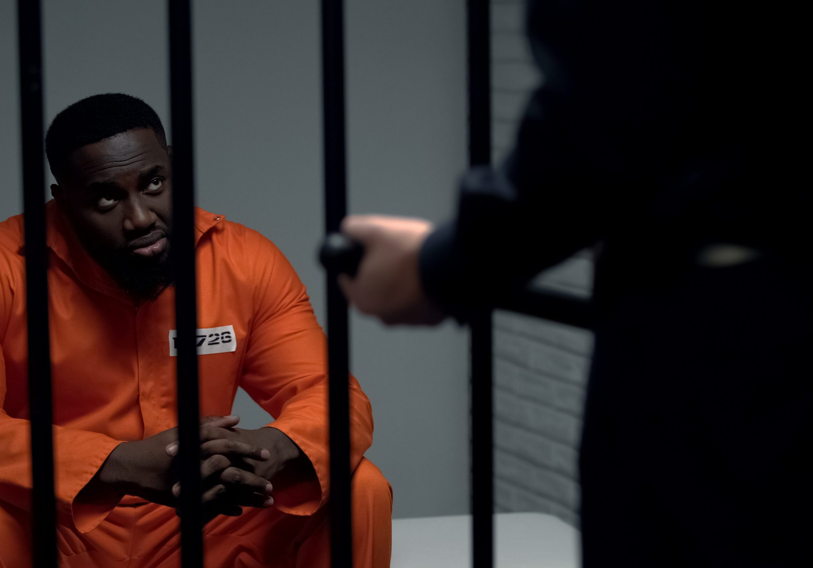 Jail guard with baton looking at afro-american prisoner in cell, harassment