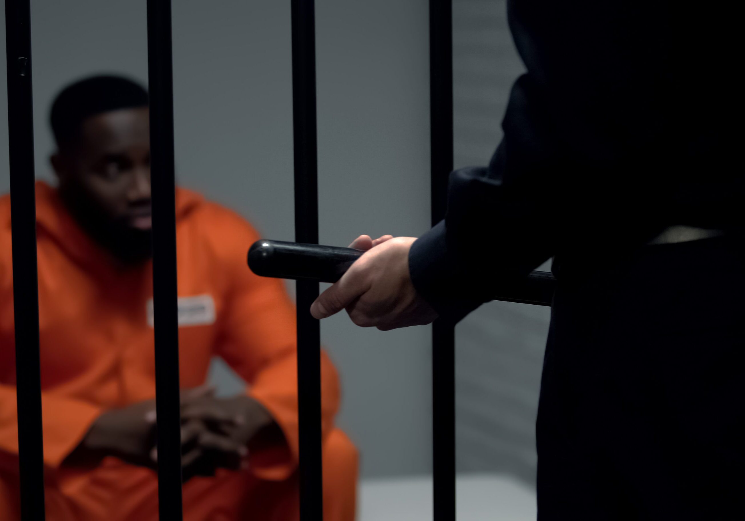 Afro-american prisoner in cell looking at jail guard with baton, harassment