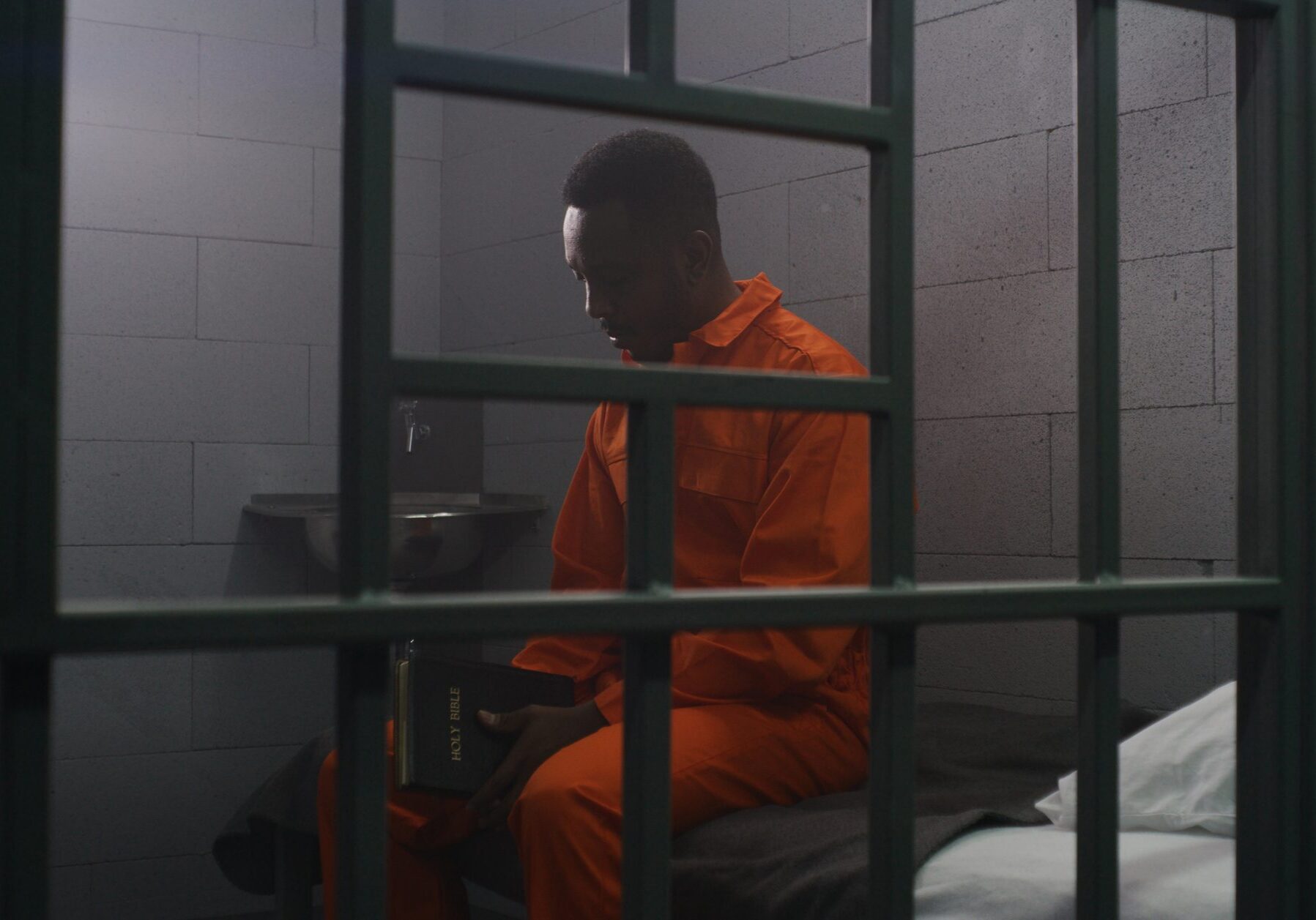 African American prisoner in orange uniform sits on the bed behind bars, reads Bible in prison cell. Criminal serves imprisonment term for crime in jail. Detention center or correctional facility.