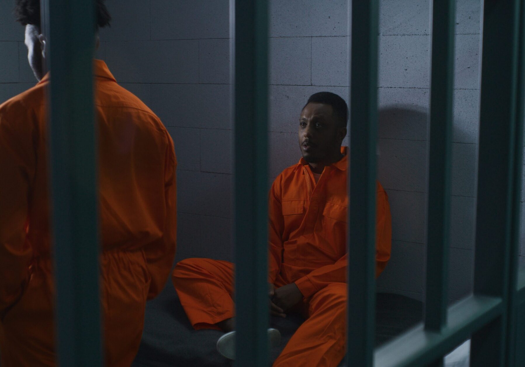 Two African American prisoners in orange uniforms talk. One man sits on bed, another walks in prison cell. Men serve imprisonment terms for crimes in jail, detention center or correctional facility.