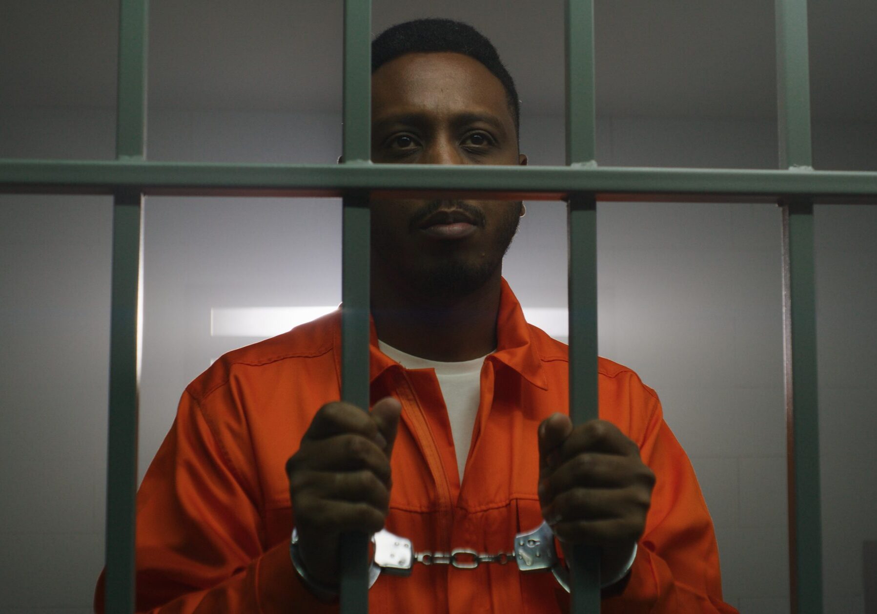 African American prisoner in orange uniform keeps hands in handcuffs on jail cell bars and looks at camera. Guilty criminal serves imprisonment term in prison. Sad murderer in correctional facility.