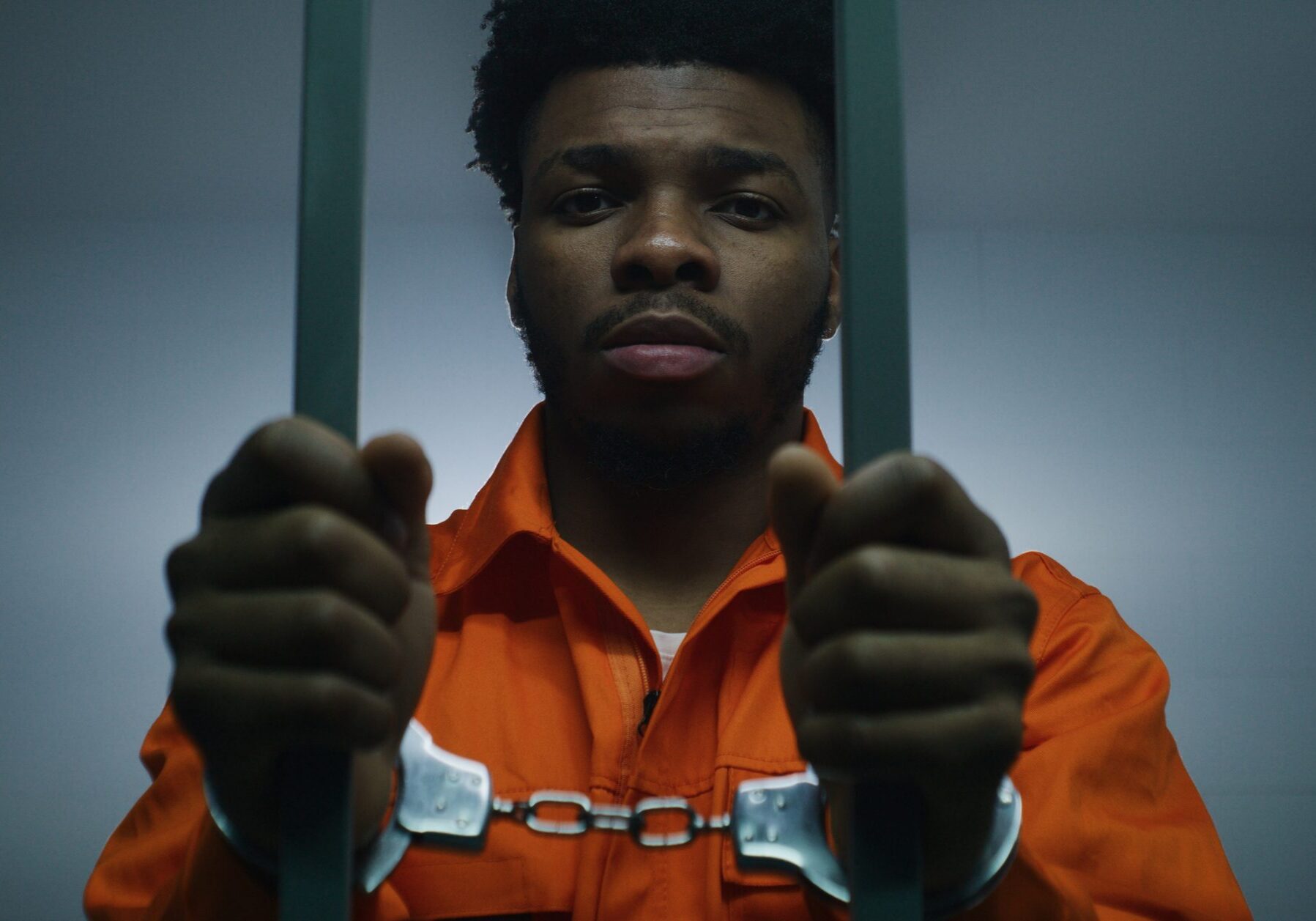 African American man in orange uniform keeps hands in handcuffs on jail cell bars. Depressed murderer serves imprisonment term in prison. Guilty prisoner in correctional facility or detention center.
