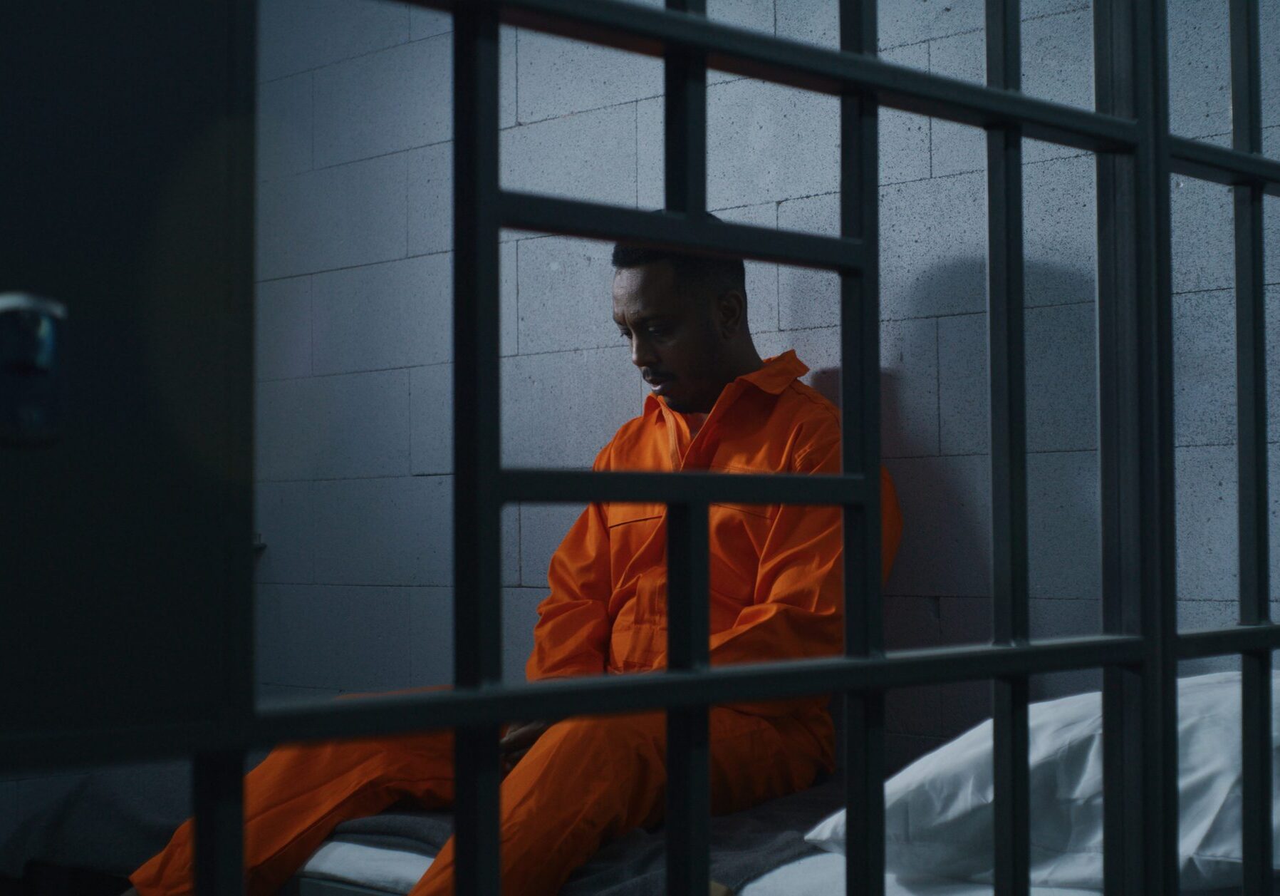 Guilty African American man in orange uniform sits on prison bed and thinks about freedom. Gloomy criminal in correctional facility or detention center. Prisoner serves imprisonment term in jail cell.