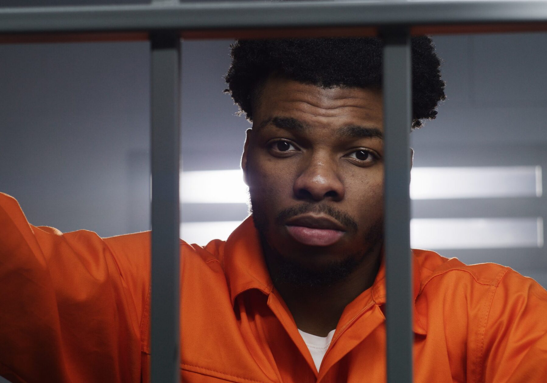 African American man in orange uniform looks at camera and kneads neck in prison cell. Prisoner serves imprisonment term in jail. Criminal in correctional facility or detention center. Portrait view.
