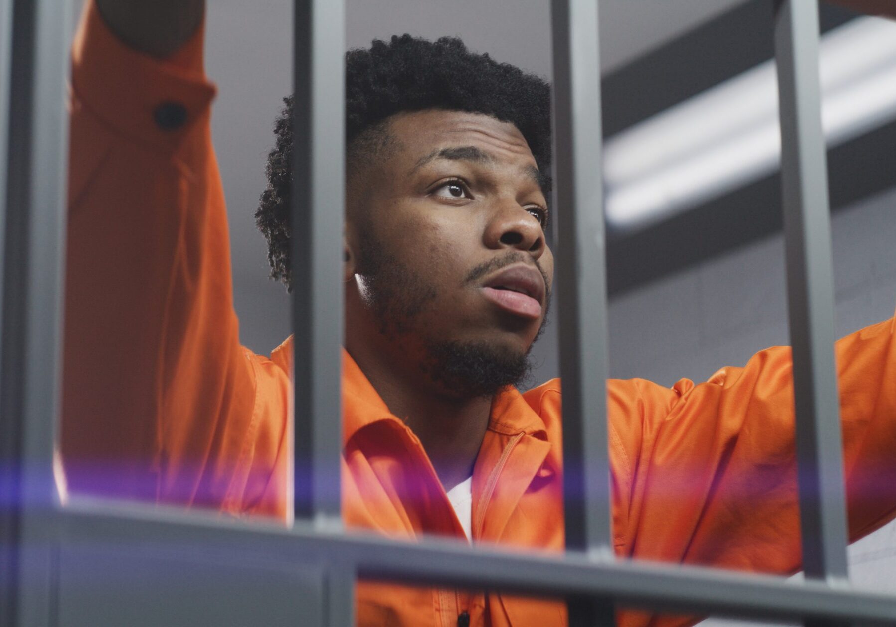 African American man in orange uniform leans hands on prison cell bars and kneads neck. Depressed prisoner serves imprisonment term in jail. Murderer in correctional facility or detention center.