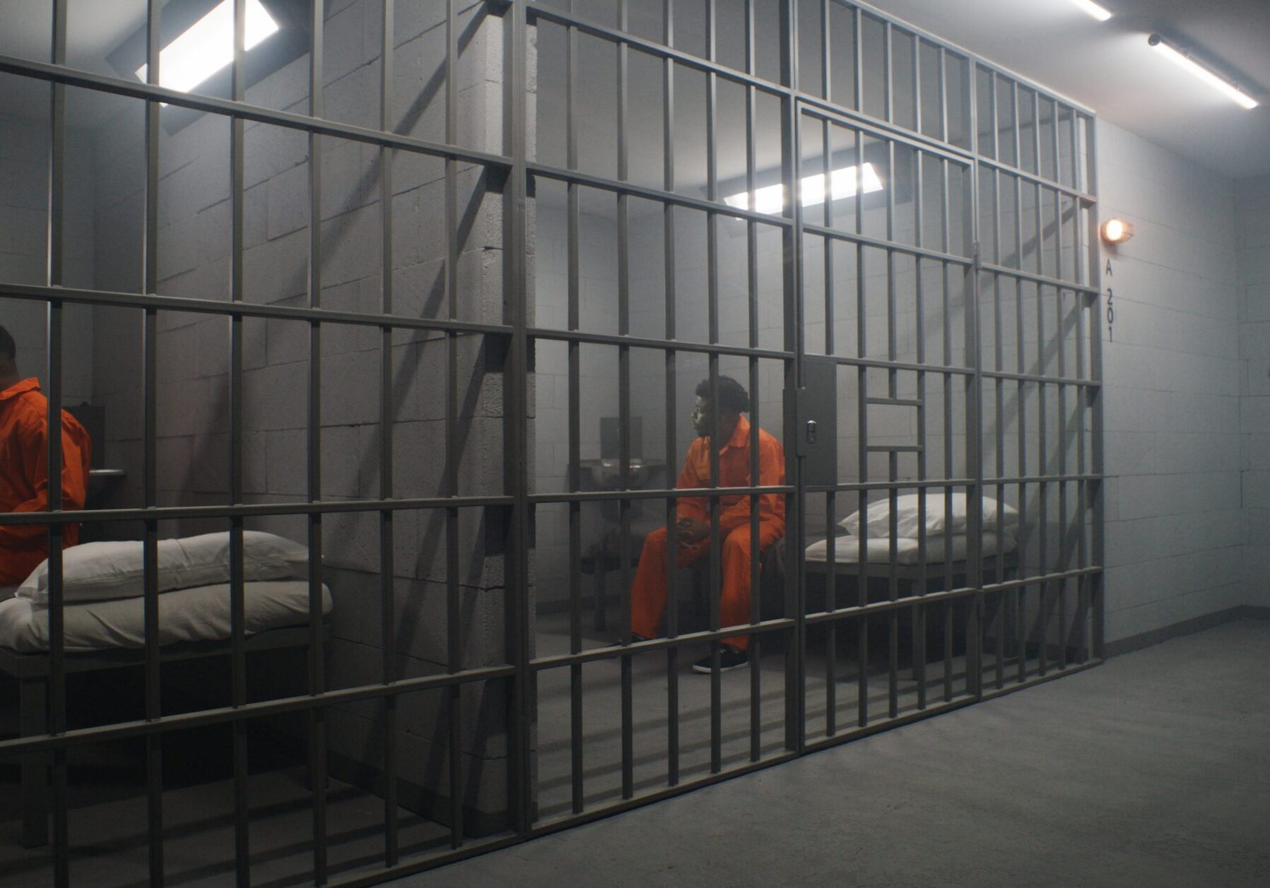 African American inmates in detention center or correctional facility. Depressed man in orange uniform sits on prison bed and looks at barred window. Prisoners serve imprisonment term in jail cell.