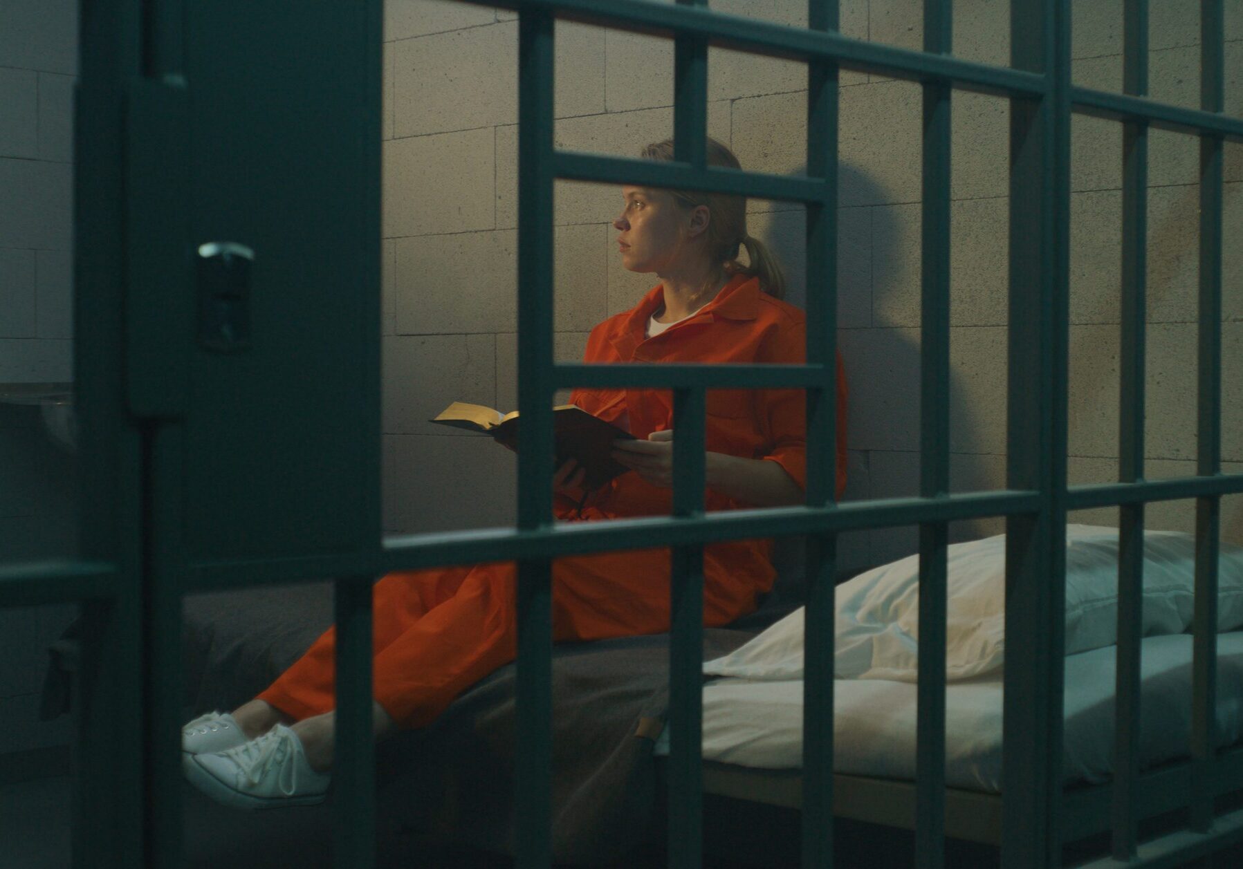 Female prisoner in orange uniform sits on bed behind metal bars, reads Bible in prison cell, looks at barred window. Woman criminal serves imprisonment term for crime in jail or correctional facility.