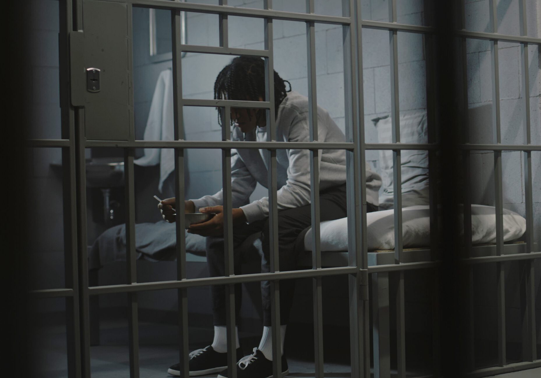 African American young prisoner eats disgusting prison food from iron bowl sitting on bed in prison cell. Teenage criminal, inmate serves imprisonment term for crime in jail. Youth detention center.