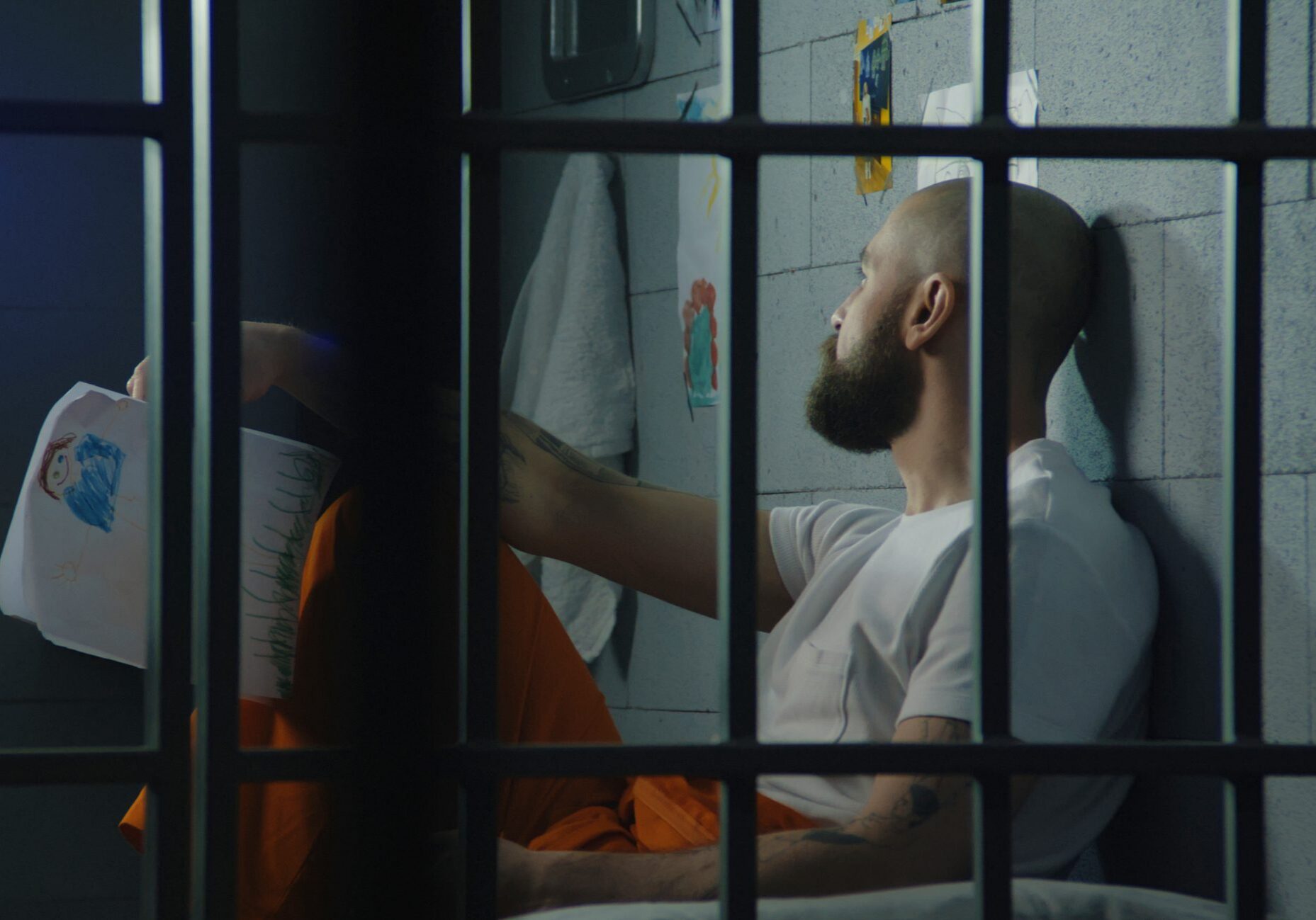 Depressed male prisoner holds drawings of child and looks at window sitting on bed in jail cell. Illegally convicted man serves imprisonment term in prison. Detention center or correctional facility.